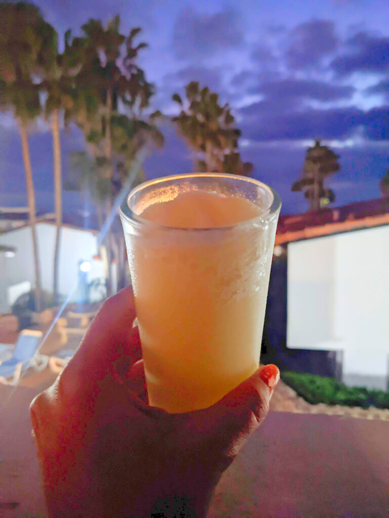 A hand holding a glass of non-alcoholic pina colada drink, with palm trees, condos and the sky in the background.
